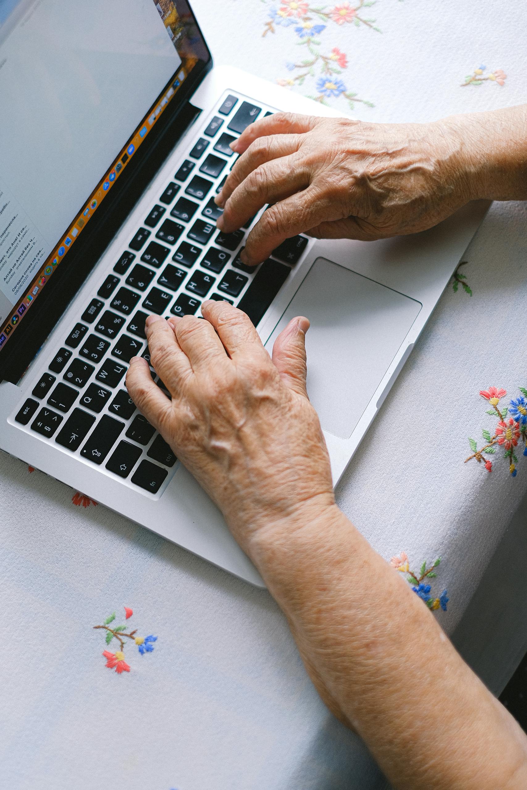 Elderly hands typing on a laptop keyboard at home, promoting digital literacy among senior adults.